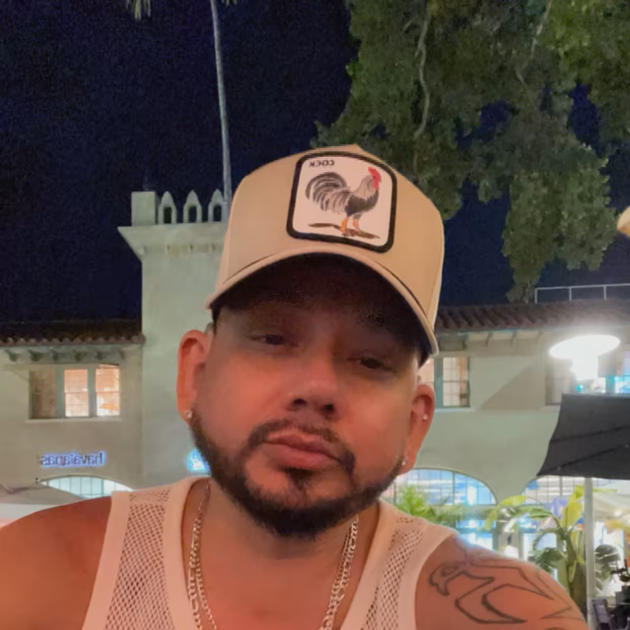 Latino man with trimmed beard and mustache, beige cap with rooster patch, white mesh tank, silver chain, outdoors at night.